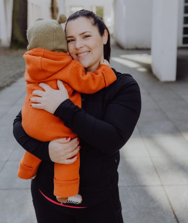 Verena, Inhaberin von Tragemamas Verena trägt ihr kleines Kind in orangener Kleidung und lächelnd auf dem Arm.